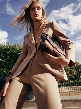 Fashion editorial shot: A blonde adult model in a tan suit holds a structured leather handbag against a sky and tree background.