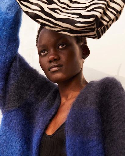 Close-up of a young adult woman with short dark hair, wearing a dark top and an open blue cardigan, with a black and white head wrap.