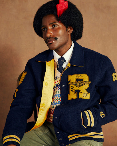 Man wearing a blue and yellow Polo letterman jacket in front of a brown background, posing with a serious expression and 1970s afro.