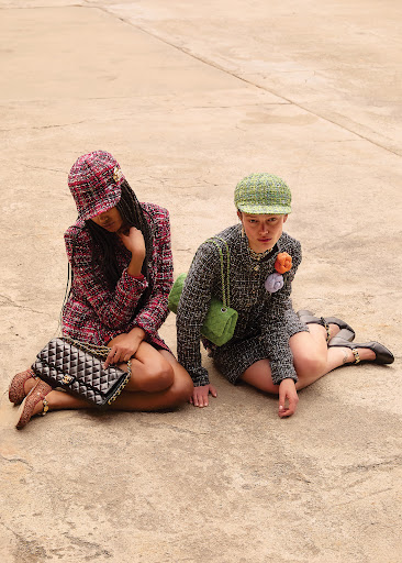 Two women in tweed outfits sit outdoors on a concrete surface wearing matching hats and purses. One is holding a Chanel handbag.