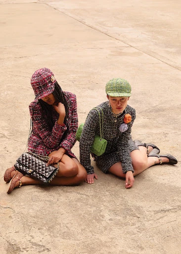 Two women in tweed outfits sit outdoors on a concrete surface wearing matching hats and purses. One is holding a Chanel handbag.