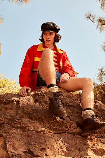 Adult male model posing on a rocky landscape wearing a red jacket, striped shirt, jean shorts, black cap, and hiking boots in an outdoor setting.