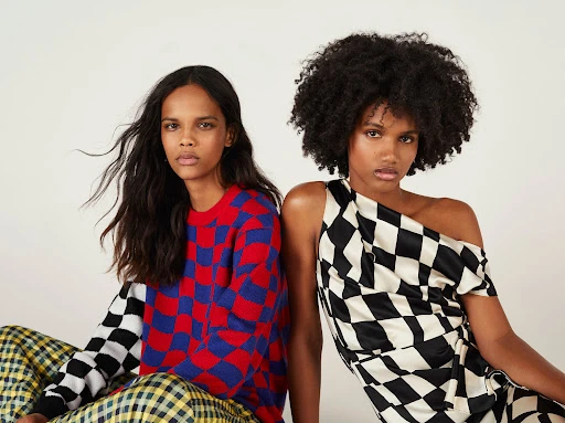 Two dark-skinned adult fashion models wearing vibrant checkered print clothing, posing in front of a clean white backdrop in a studio setting.