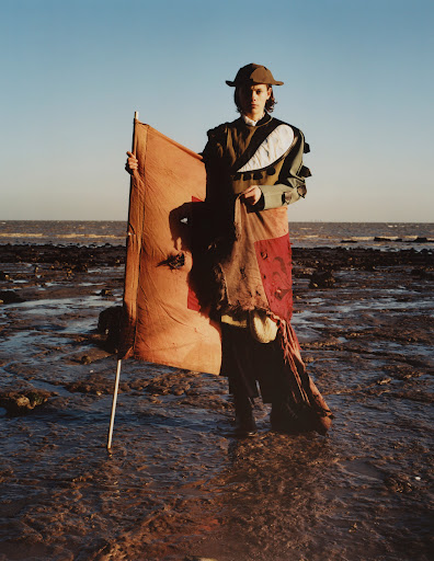 A person stands on a rocky beach holding a tan-colored flag against a clear blue sky and distant ocean, wearing an olive green outfit and a hat.
