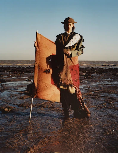 A person stands on a rocky beach holding a tan-colored flag against a clear blue sky and distant ocean, wearing an olive green outfit and a hat.