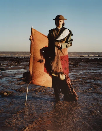 A person stands on a rocky beach holding a tan-colored flag against a clear blue sky and distant ocean, wearing an olive green outfit and a hat.