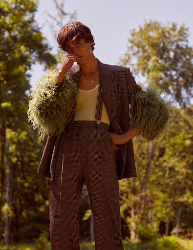 Young adult poses outdoors in a brown suit with feathered sleeves, captured in natural light against a backdrop of green foliage.