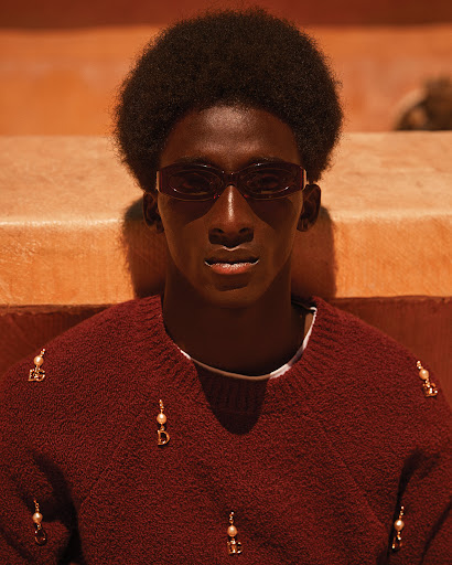 Portrait of African American male model with sunglasses wearing an embellished sweater against a minimalist orange background.