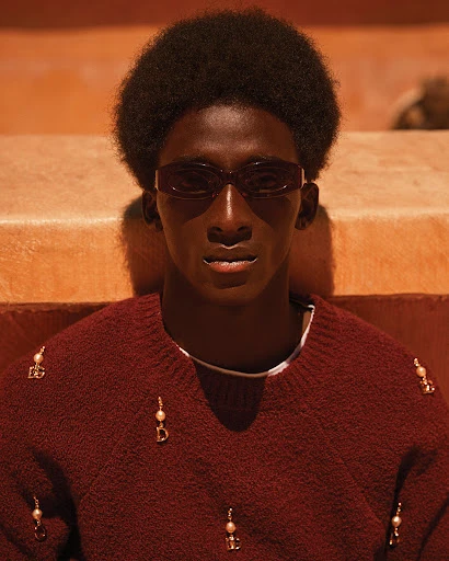Portrait of African American male model with sunglasses wearing an embellished sweater against a minimalist orange background.
