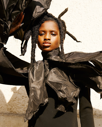 Portrait of an African woman with locs wearing a black garment with exaggerated sleeves against a solid, neutral background.