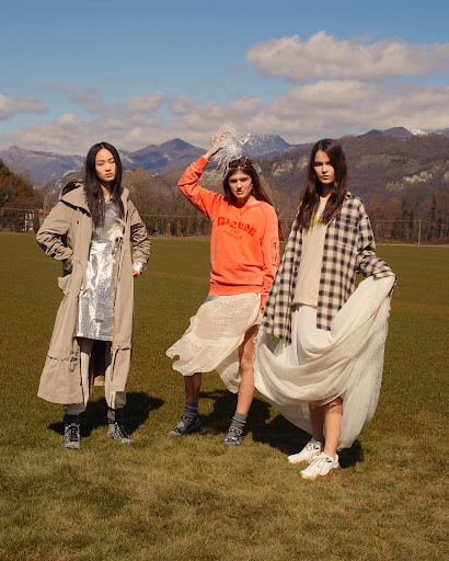 Three women pose in stylish outfits outdoors with mountains in background. One wears a beige trench coat, one wears an orange sweater, one a plaid shirt.
