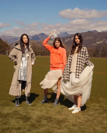 Three women pose in stylish outfits outdoors with mountains in background. One wears a beige trench coat, one wears an orange sweater, one a plaid shirt.