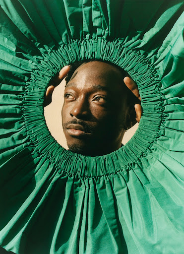 Close-up studio portrait of a Black male model framed by a large green ruffled collar, hands near his face, and looking off to the side.