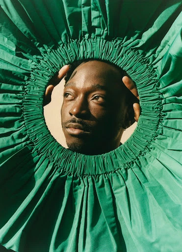 Close-up studio portrait of a Black male model framed by a large green ruffled collar, hands near his face, and looking off to the side.
