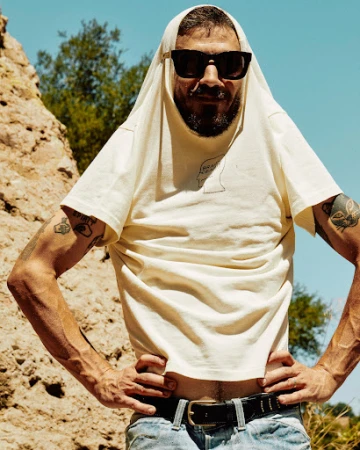 Man wearing sunglasses with a white t-shirt pulled over his head standing outdoors against a rocky terrain background