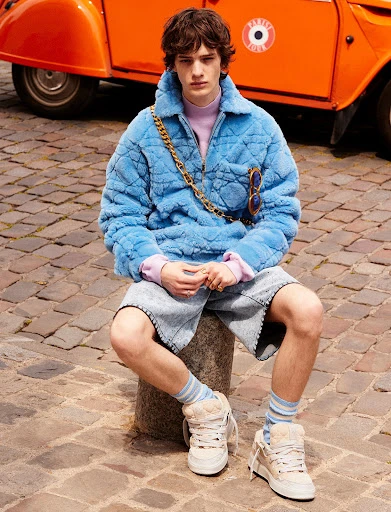 Young man poses on cobblestone street with orange car in background, wearing blue faux-fur jacket, lilac turtleneck, jean shorts, socks, and sneakers.
