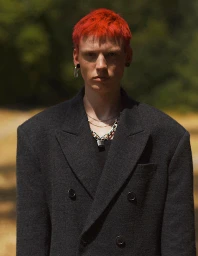 Portrait of a man with bright red hair wearing a dark grey oversized blazer and layered necklaces against an outdoor background