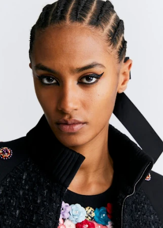 Close up portrait of woman with cornrows and dramatic winged eyeliner wearing a black tweed jacket with colorful floral embroidery
