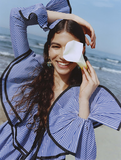 Smiling woman with dark hair poses on beach wearing a striped top, holding a white lily in front of one eye in an outdoor shot.