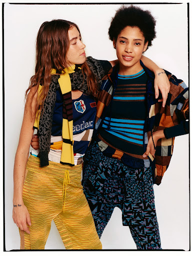Two young adult women pose in a brightly lit photo studio wearing patterned tops and bottoms, accessorized with a scarf and jacket.