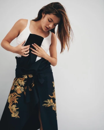 Woman in a white tank top and dark skirt with gold embroidery against a light background