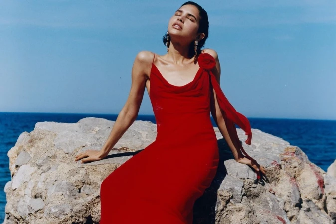 Woman in a red slip dress seated on a coastal rock formation against an ocean backdrop under bright sunlight.