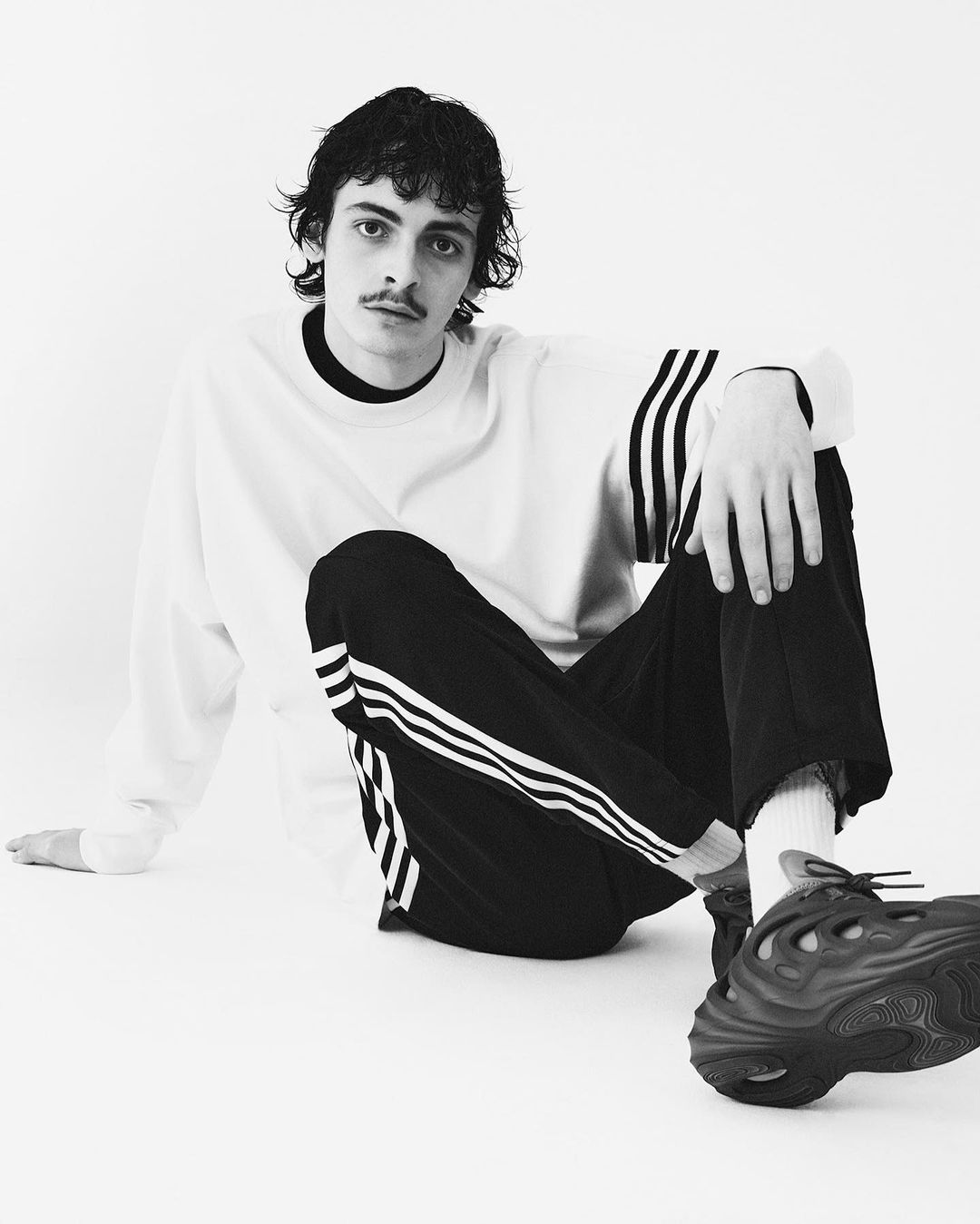 Black-and-white portrait of a young adult male with a mustache, wearing a white sweatshirt and striped black pants, seated against a white background.