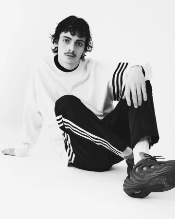 Black-and-white portrait of a young adult male with a mustache, wearing a white sweatshirt and striped black pants, seated against a white background.