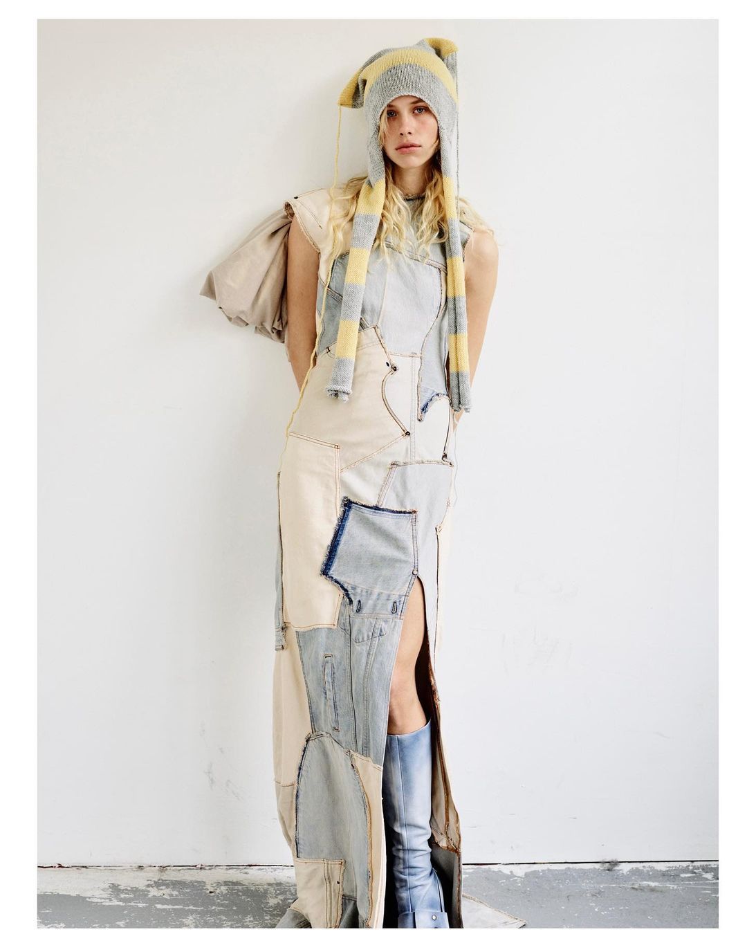 A model poses in a patchwork denim dress with a thigh-high slit, paired with a striped knit hat, against a white wall in a studio setting.