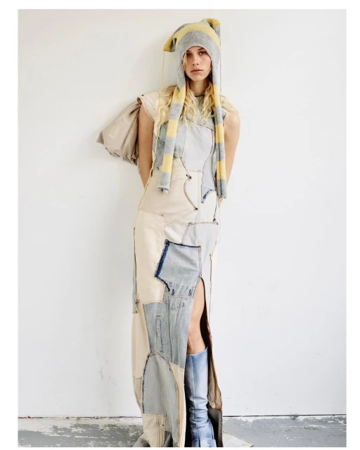 A model poses in a patchwork denim dress with a thigh-high slit, paired with a striped knit hat, against a white wall in a studio setting.