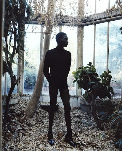 Dark-skinned adult poses in a greenhouse with dark clothing, surrounded by green plants and fallen leaves, captured in medium shot.