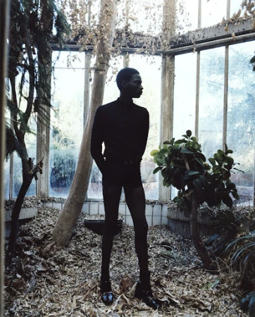 Dark-skinned adult poses in a greenhouse with dark clothing, surrounded by green plants and fallen leaves, captured in medium shot.