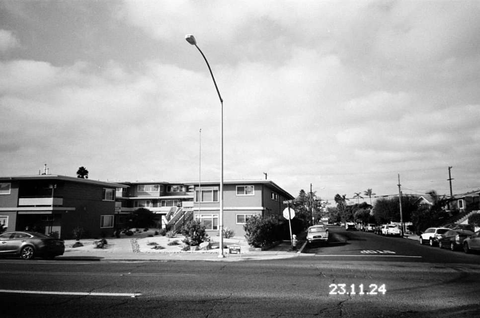 Black and white photograph of a street in a residential neighborhood with apartment buildings and parked cars under a cloudy sky.