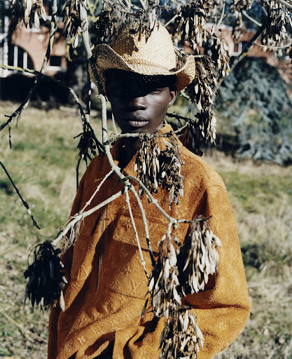 Portrait of an African male in an orange top and straw hat, standing in a grassy field, framed by tree branches with dried leaves.