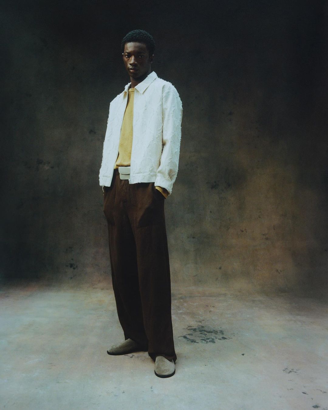A full shot of a Black male model in a cream jacket, brown pants, and brown shoes, posed against a mottled brown backdrop in a studio.