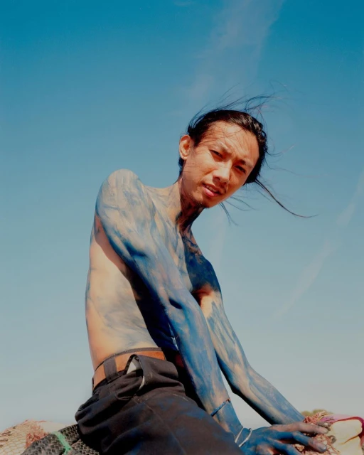 Young adult male with blue body paint sitting outdoors against a clear blue sky in a medium portrait shot. Windblown hair.