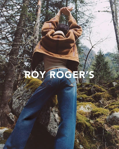 A person standing among trees and mossy rocks, wearing denim and a brown jacket, with a text overlay of ROY ROGER'S.