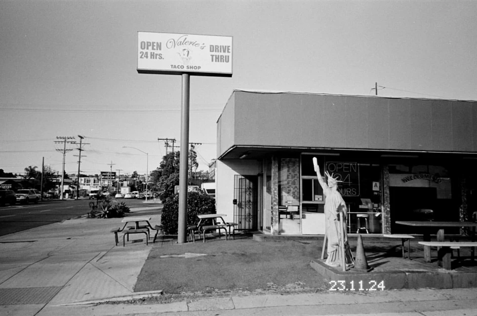 Retro black and white outdoor view of Valerie's Drive Thru taco shop featuring a Statue of Liberty replica and picnic tables.