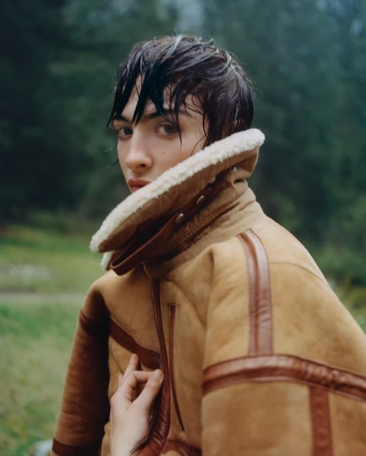 Portrait of a person with wet hair wearing a brown shearling jacket standing in a forest setting on an overcast day.