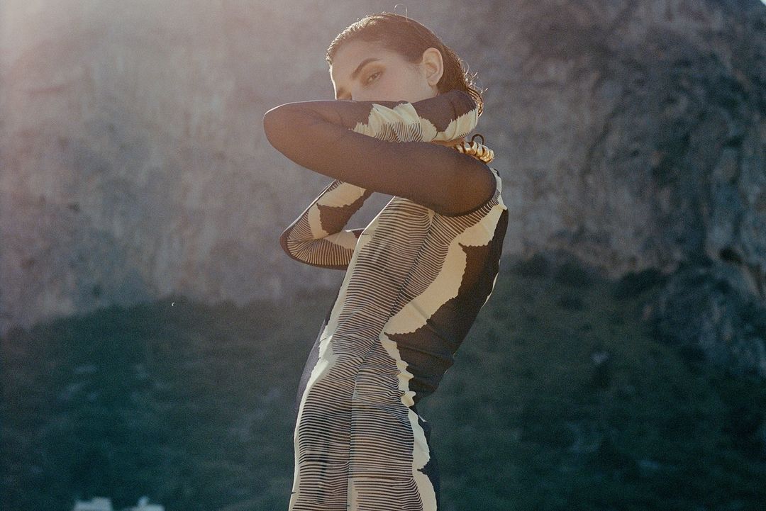 Adult female fashion model strikes a pose outdoors in a textured bodycon dress with a high mountain visible in the background.