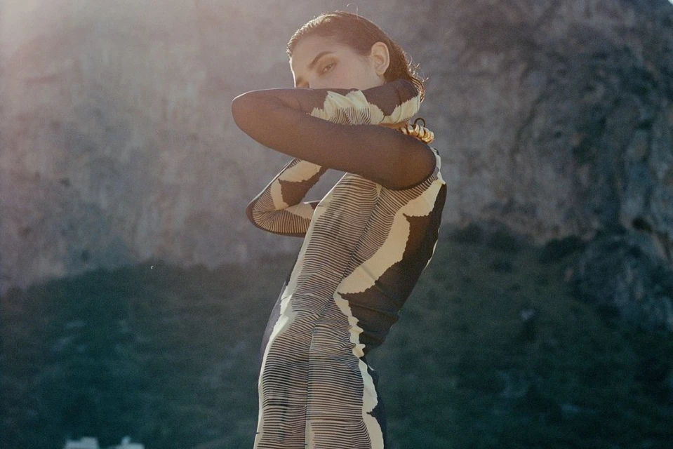 Adult female fashion model strikes a pose outdoors in a textured bodycon dress with a high mountain visible in the background.