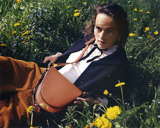 Woman lies in a field of dandelions, wearing a navy jacket and carrying a brown leather shoulder bag. Corduroy pants are visible.