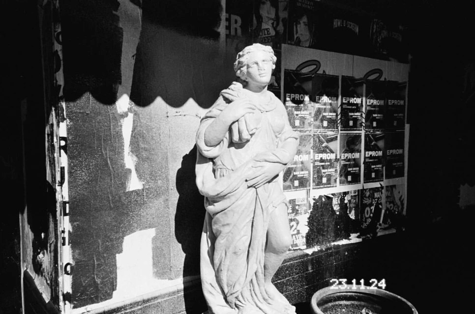 Monochrome photograph of a classical statue in front of a wall covered in posters, next to a trash can in an indoor setting.