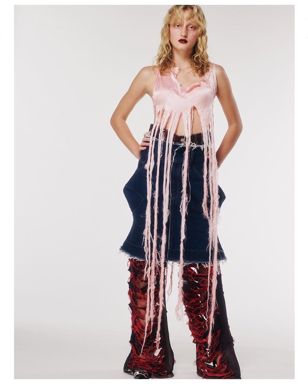 Young woman in a pink fringed top and deconstructed denim skirt with torn red and black leggings poses in a photo studio.