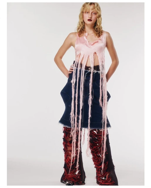 Young woman in a pink fringed top and deconstructed denim skirt with torn red and black leggings poses in a photo studio.