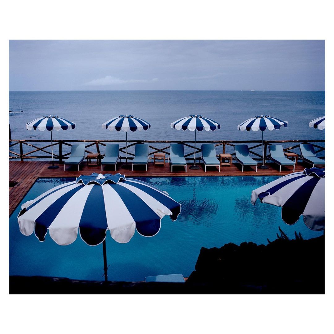 View of a pool surrounded by blue and white striped umbrellas and lounge chairs, with the ocean in the background.