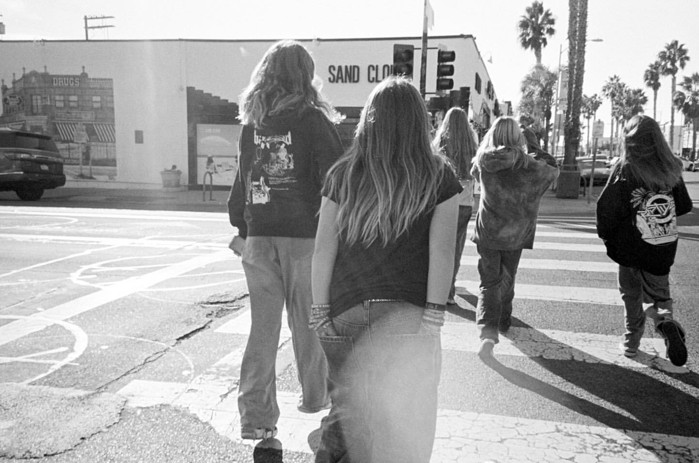 Black and white street scene with five young people walking on a crosswalk, palm trees and buildings in the background, sunny day.