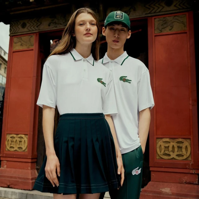 A young woman and man in matching white Lacoste polo shirts and dark green bottoms stand in front of an ornate, red building exterior.