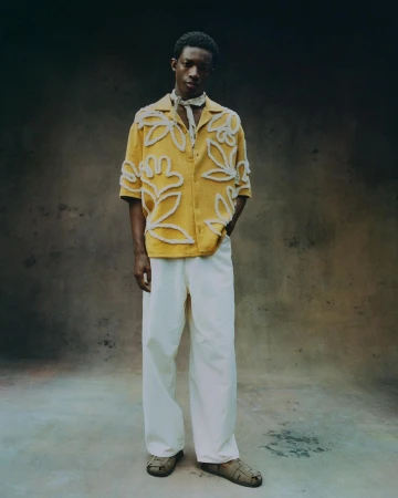 Man wearing a yellow embroidered short-sleeve shirt and white trousers standing against a textured studio backdrop