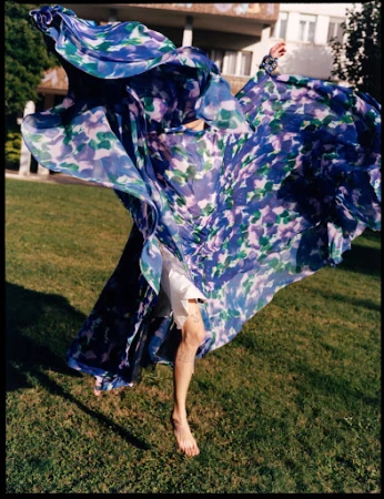 Model in a billowing blue and purple floral dress leaping above a green grass lawn with a building facade behind